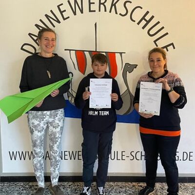 (von links) Schulleiterin A. Schönberg mit Schüler und Lehrkraft D. von Klein vor dem Logo der Dannewerkschule-Schleswig